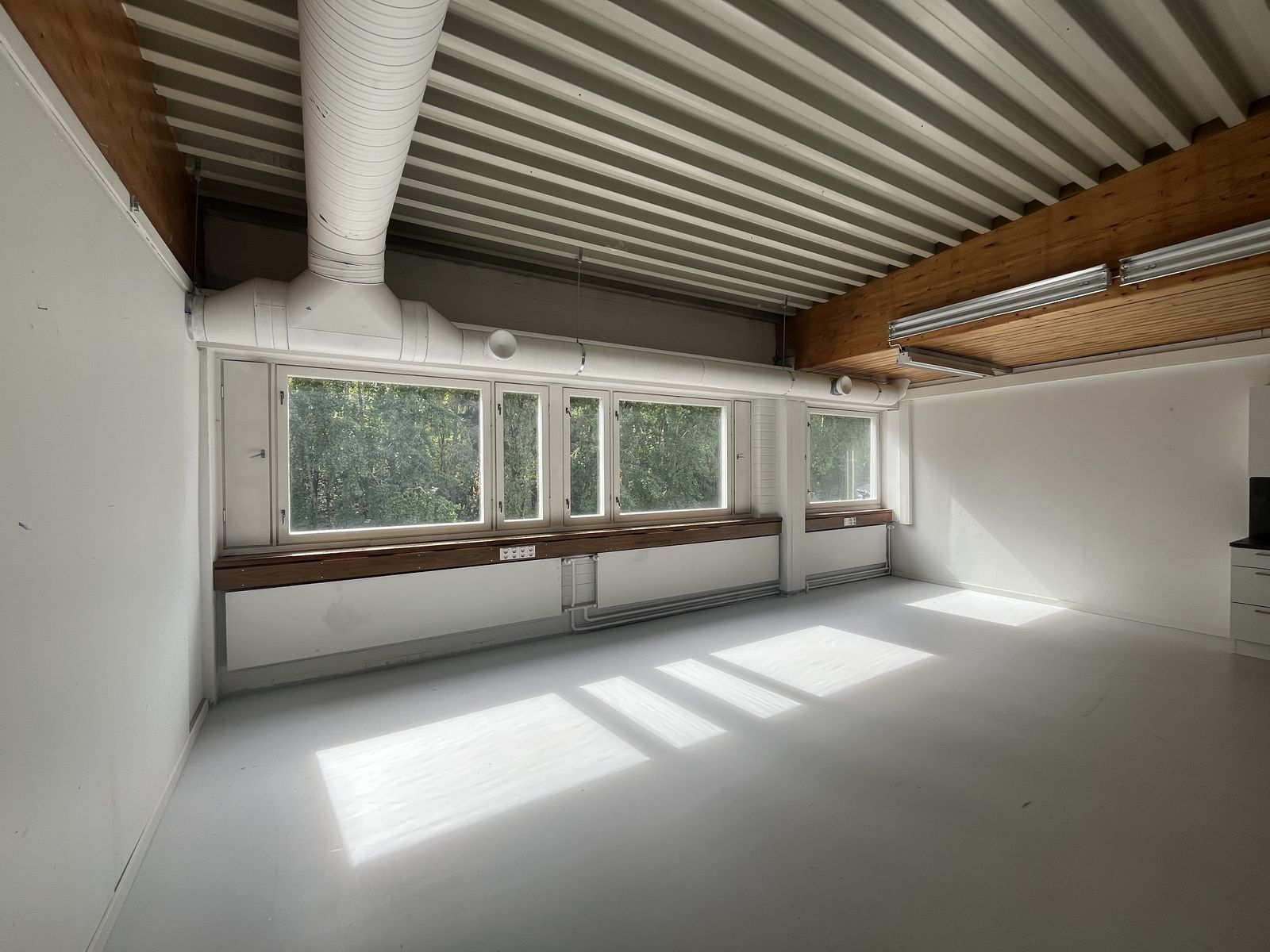The BLAZE studio: an empty white room with a row of windows looking out onto birch trees, a corrugated ceiling and exposed ventilation ducts.